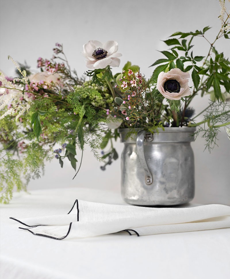 Eine altmodische metallene Vase mit Schnittblumen steht auf einem kleinen Tisch mit weißer Tischdecke, auf dem außerdem eine weiße Stoffserviette mit schwarzem Saum liegt