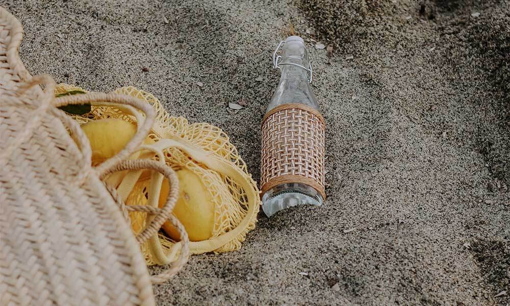 Eine Basttasche mit Zitronen liegt neben einer Glasflasche im Sand