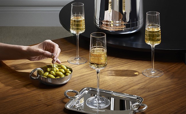 Drei gefüllte Champagnergläser stehen auf einem Holztisch - eines davon auf einem silbernen Tablett - während sich eine Hand eine Olive aus einer silbernen Schüssel aufspießt - Im Hintergrund steht ein rundes, schwarzes Tablett mit einem silbernen Champag