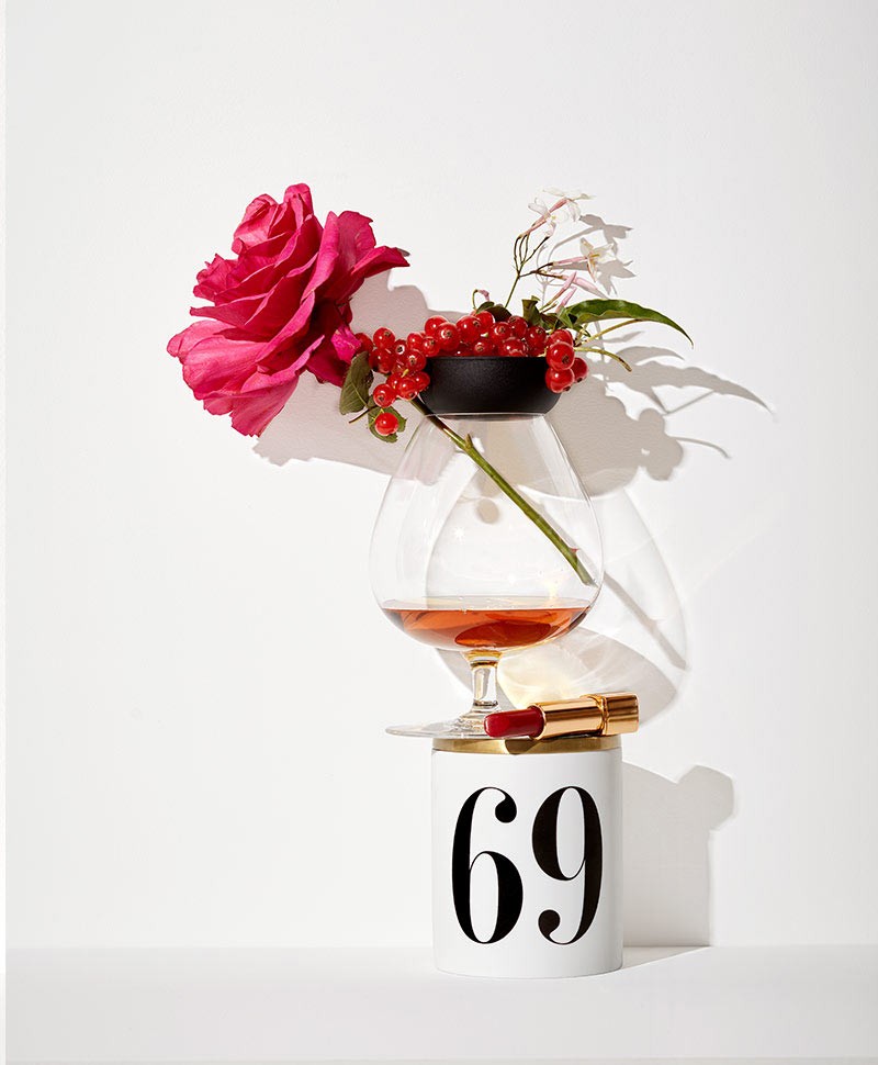 Eine Duftkerze von L'Objekt mit der Zahl 69 steht vor einem weißen Hintergrund, auf ihr ist ein Weinglas mit einer Rose, sowie ein roter Lippenstift platziert