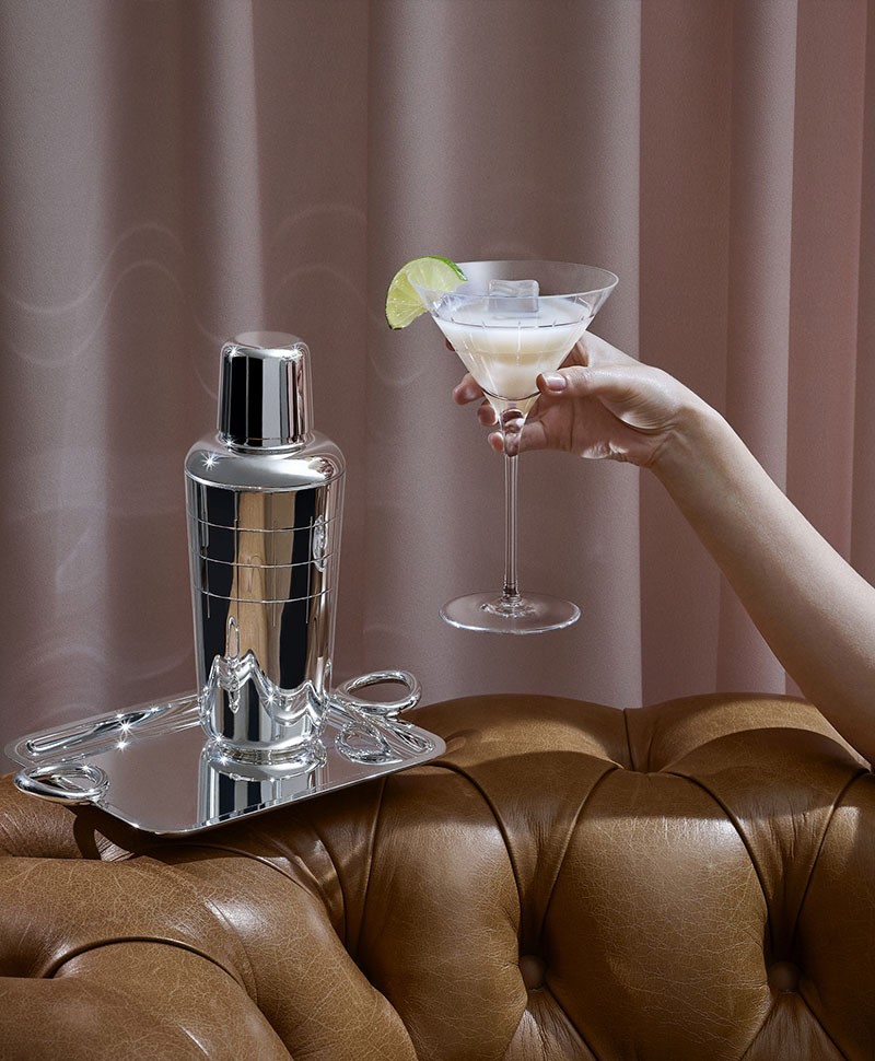 Eine Hand hält ein trichterförmiges Cocktailglas - auf einer braunen Ledercouch steht ein silbernes Tablett mit einem silbernen Cocktailshaker