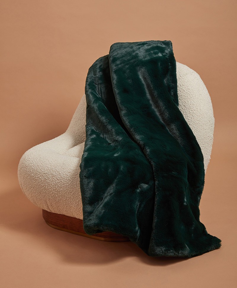 Das Moodbild zeigt die Decke Brady aus Kunstfell von der Marke Apparis in der Farbe emerald green auf einem weißen Sessel – im Onlineshop RAUM concept store