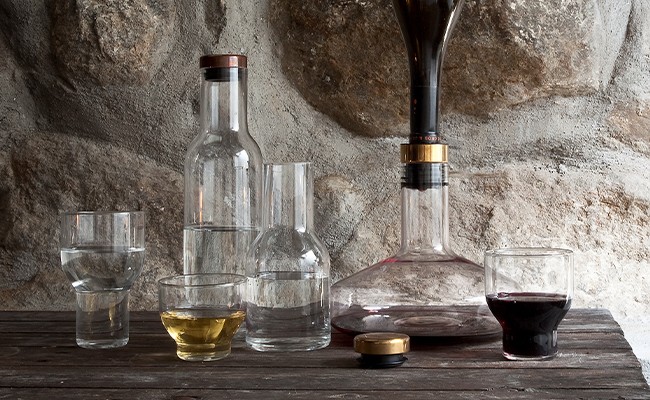 Weindekanter, Karaffen und zum Teil halb gefüllte Gläser stehen auf einer dunklen Holzoberfläche vor einem steinernen Hintergrund