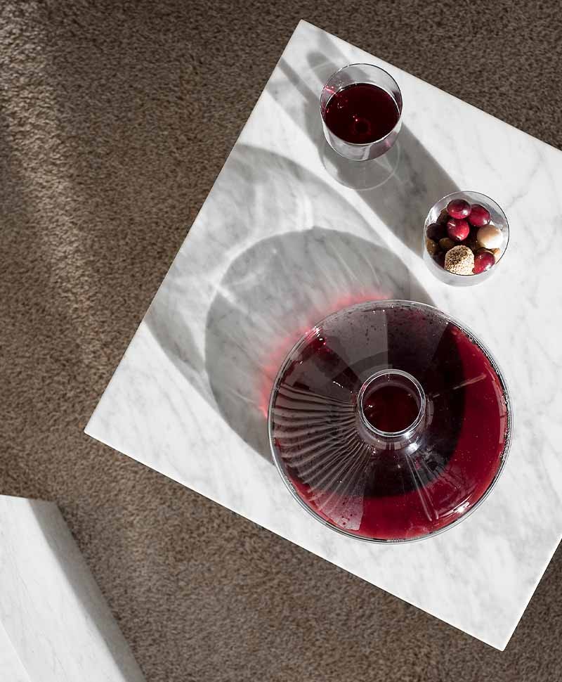 Ein geöffneter Weindekanter mit Inhalt steht zusammen mit einem gefülltem Weinglas und einer kleinen Dessertschale mit Früchten auf einer quadratischen Marmorplatte