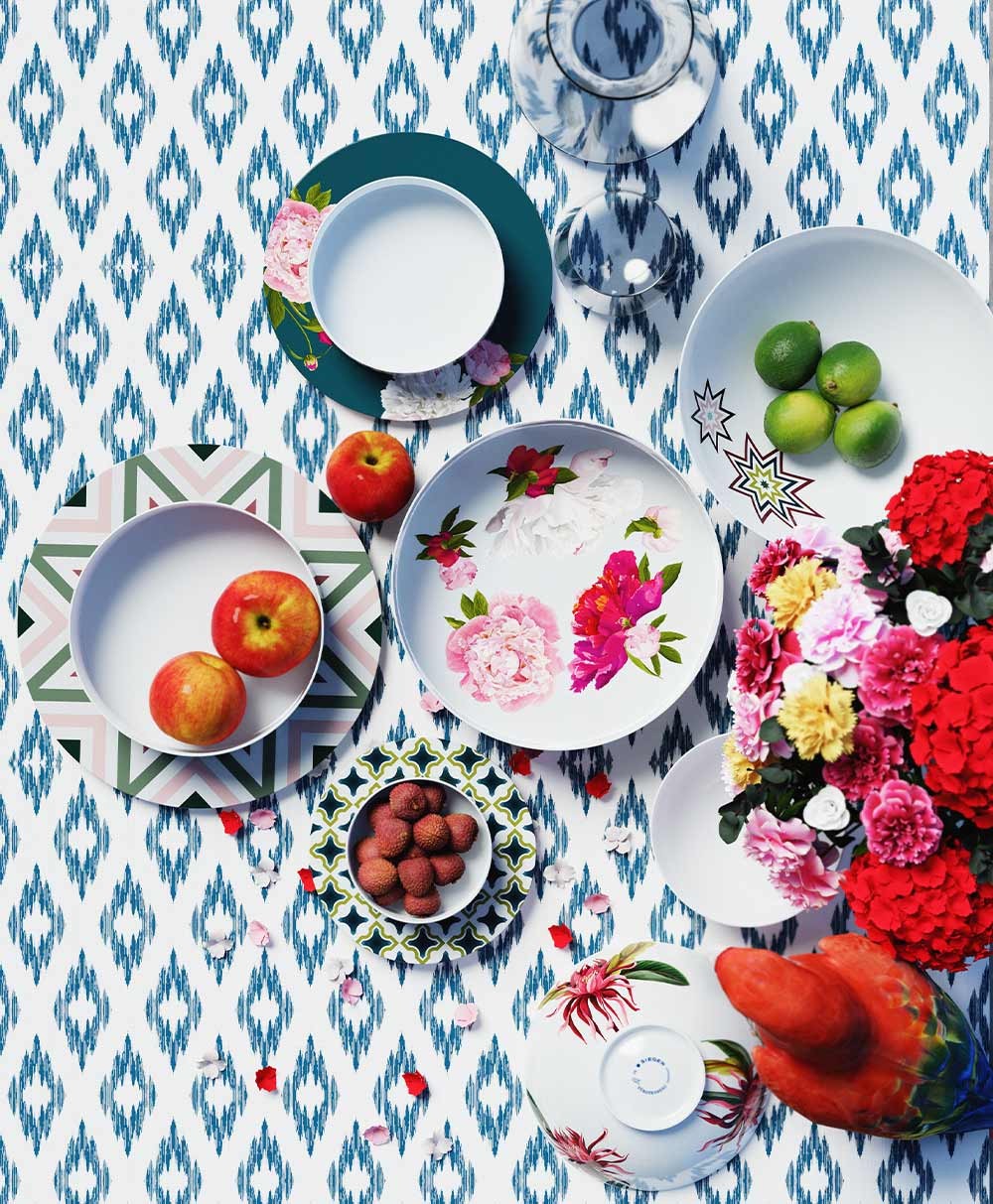 Moodbild, das einige Teller und Schalen der Paraiso-Serie von Sieger by Fürstenberg zeigt, die dekorativ mit Blumen und Obst auf einer blau. gemusterten Unterlage arrangiert wurden