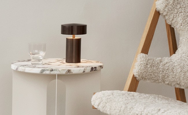 Die Column Table Lampe von Menu Design steht auf einem kleinen Marmor-Beistelltisch, daneben steht ein niedriger Holzstuhl mit weißem Polster