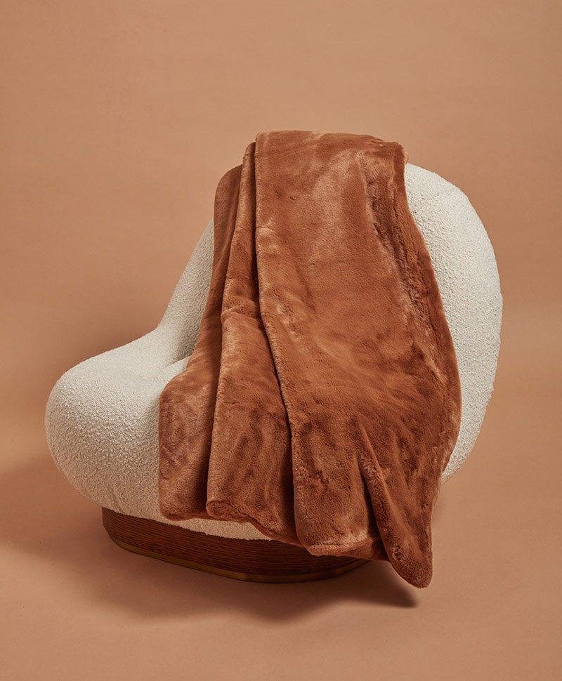 Das Moodbild zeigt die Decke Brady aus Kunstfell von der Marke Apparis in der Farbe camel auf einem weißen Sessel – im Onlineshop RAUM concept store
