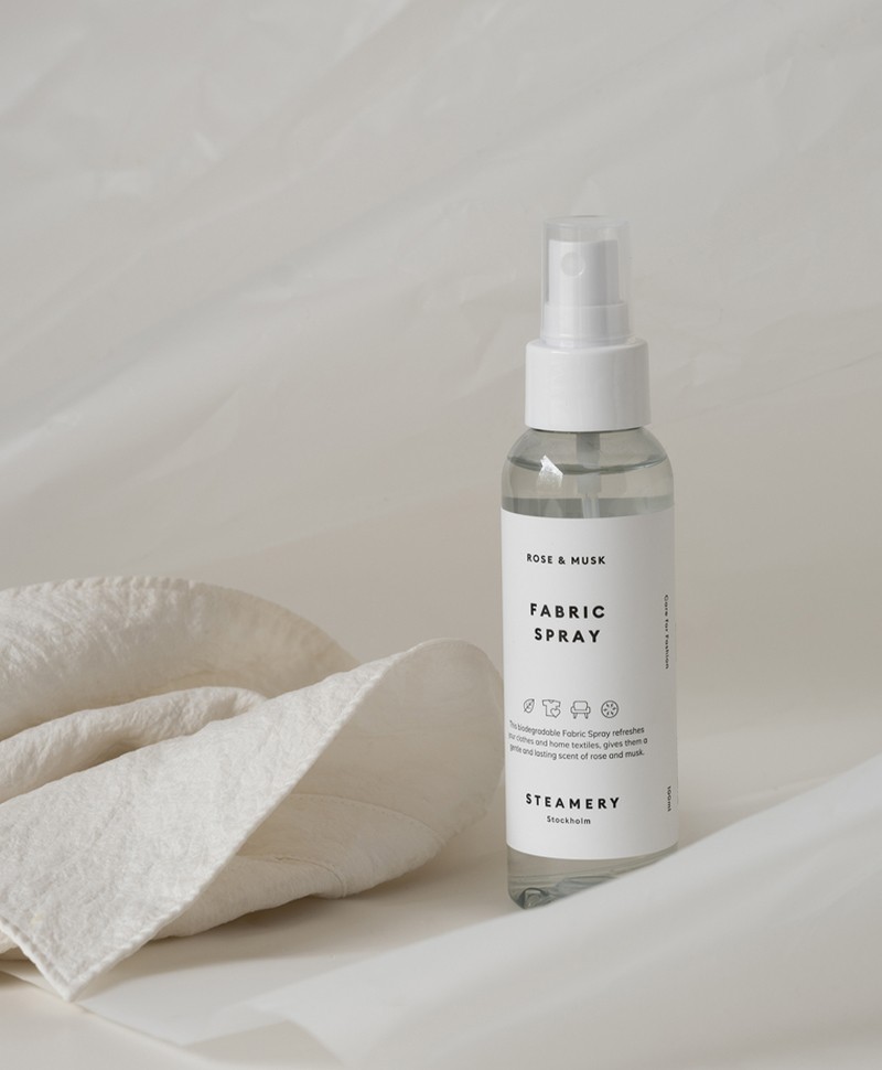 Moodbild des Fabric Sprays der Marke Steamery, das vor einem texturierten weißen Hintergrund steht - daneben liegt ein ebenfalls weißes Stofftuch